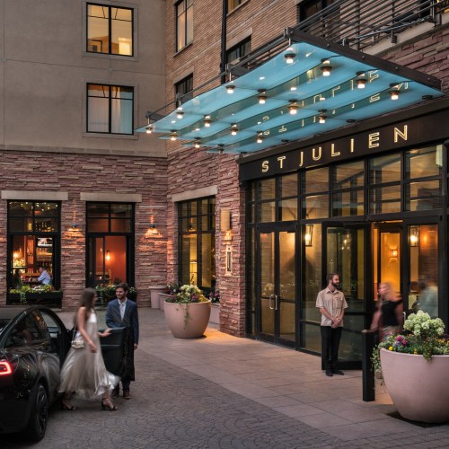 A stylish hotel entrance with a blue awning, featuring guests arriving and vibrant flower pots. Evening ambiance.