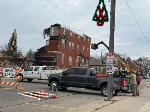 Demolition of a brick building with construction equipment; a "Road Closed" sign and holiday decoration visible.