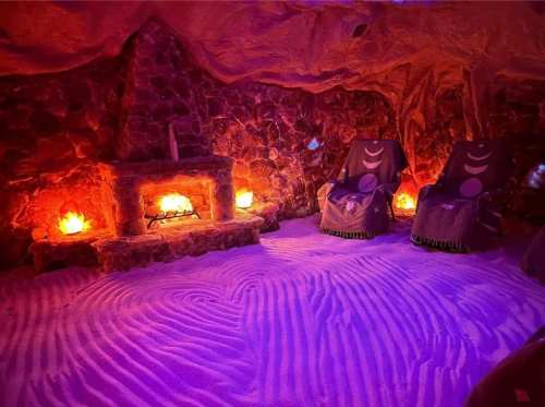 A cozy salt room with stone walls, two chairs, and glowing salt lamps, featuring a sandy floor with a wave pattern.