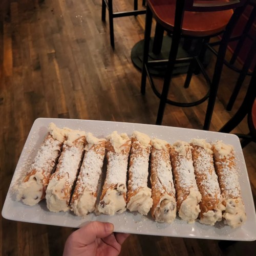 A hand holding a platter of cannoli, dusted with powdered sugar, on a wooden floor in a restaurant setting.