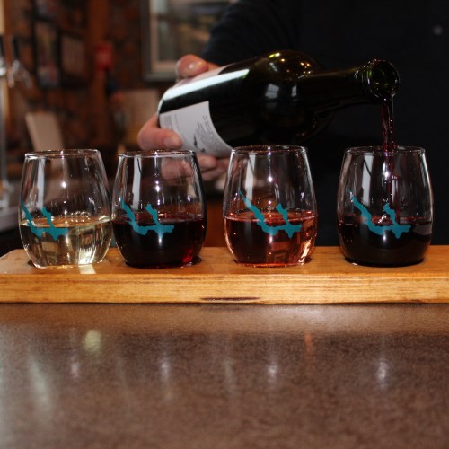 A wooden tray holds four wine glasses with different colored wines, while a person pours red wine into a glass.