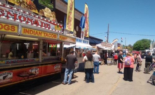 A bustling street fair with food stalls, including lemonade, hot dogs, and Indian tacos, and a crowd enjoying the event.