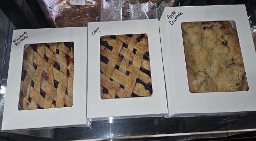 Three boxes of baked goods: a lattice-topped raspberry pie, a cherry pie, and an apple crumble.
