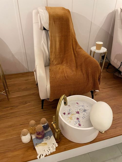 A cozy spa setup featuring a white chair, a brown blanket, and a foot bath with decorative stones and flowers.