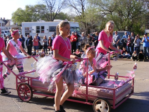 Girls in pink shirts and tutus pull a decorated wagon with a seated girl during a parade, with a crowd in the background.