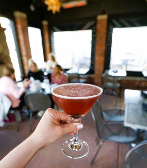 A hand holds a cocktail glass filled with a reddish drink, with a blurred background of people chatting in a cozy setting.