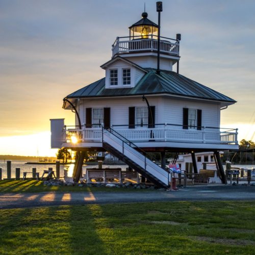 A charming lighthouse with a green roof and white exterior, illuminated by sunset, overlooking a calm waterway.