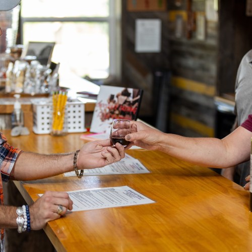 A person hands a small glass of red liquid to another at a wooden bar, with menus and drinks in the background.