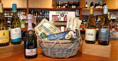 A woven basket filled with gourmet snacks, cheeses, and wines displayed on a wooden counter in a store.