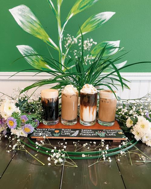 Four decorative coffee drinks on a wooden tray, surrounded by flowers and greenery against a green wall.