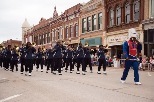 A marching band in uniforms parades down a street, led by a mascot in a colorful outfit. Crowds watch from the sidelines.