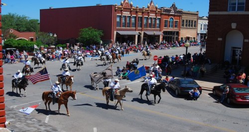 A lively parade with horseback riders, flags, and a crowd gathered along a street in a historic town.