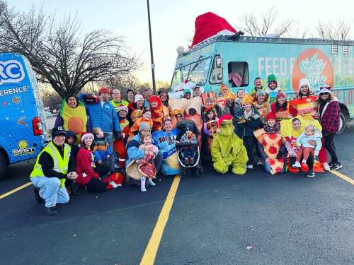 A large group of people in festive costumes poses in front of food trucks, celebrating a community event.