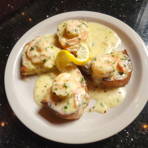 A plate of shrimp topped with creamy sauce on toasted bread, garnished with lemon and herbs.