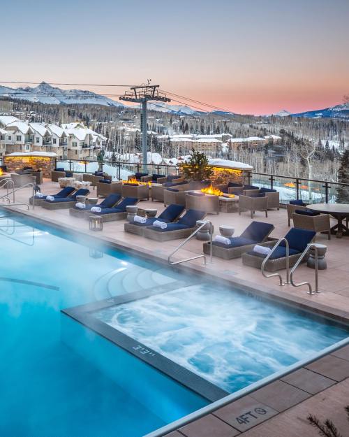 A scenic pool area with a hot tub, lounge chairs, and snowy mountains in the background at sunset.