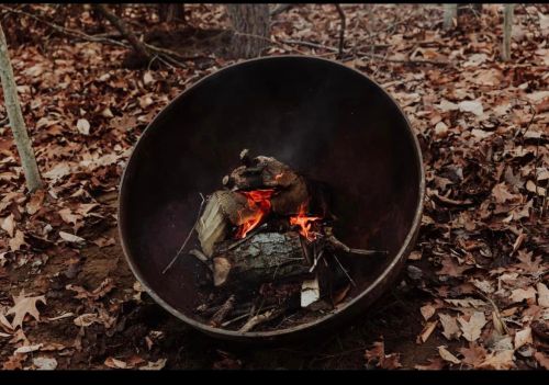 A fire burning in a large, round metal fire pit surrounded by autumn leaves in a wooded area.