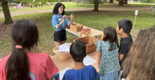 A young woman engages with children at a table with baskets in a park setting.