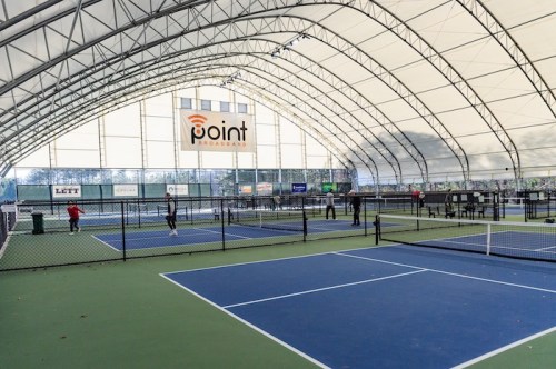 Indoor tennis facility with multiple courts, players in action, and a large "Point" sign on the wall.