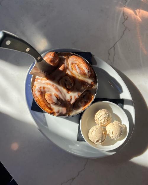 A warm cinnamon roll drizzled with icing, served in a dish alongside three scoops of vanilla ice cream.