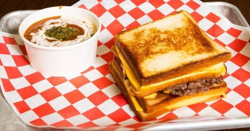 A stacked grilled cheese sandwich with melted cheese and a side of chili in a cup, served on a checkered tray.