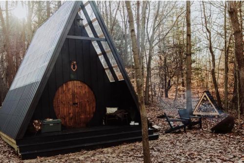 A cozy A-frame cabin in a wooded area, with a wooden door and a fire pit nearby, surrounded by fallen leaves.