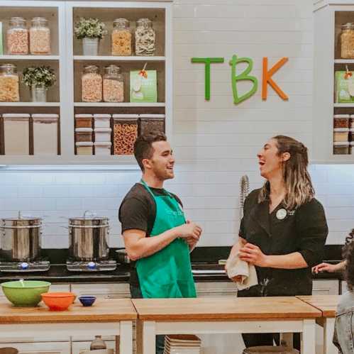 Two people in a kitchen setting, smiling and interacting, with shelves of jars and bowls in the background.