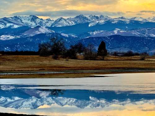 A serene landscape featuring snow-capped mountains reflected in a calm lake at sunset, with grassy fields in the foreground.