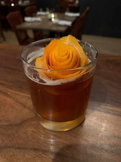A cocktail in a glass garnished with a rose made of orange peel, resting on a wooden surface.