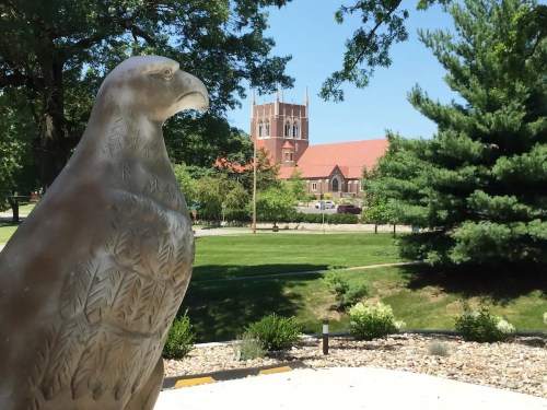 A glass eagle sculpture in the foreground with a church tower and trees in the background on a sunny day.