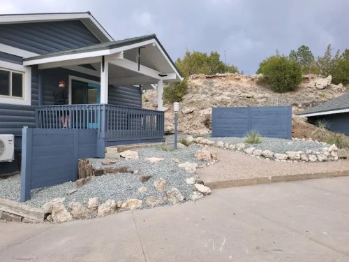 A modern blue house with a porch, surrounded by gravel landscaping and stone features, under a cloudy sky.