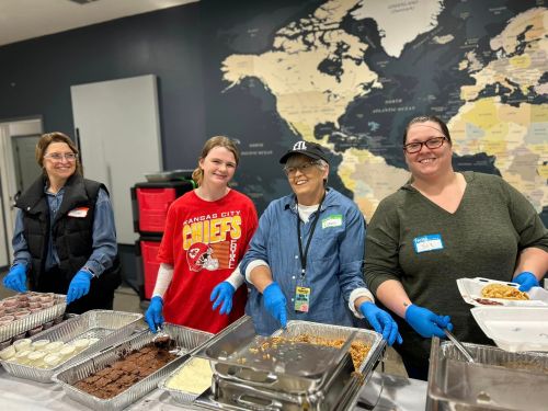Four women in gloves serve food at a community event, smiling in front of a world map backdrop.