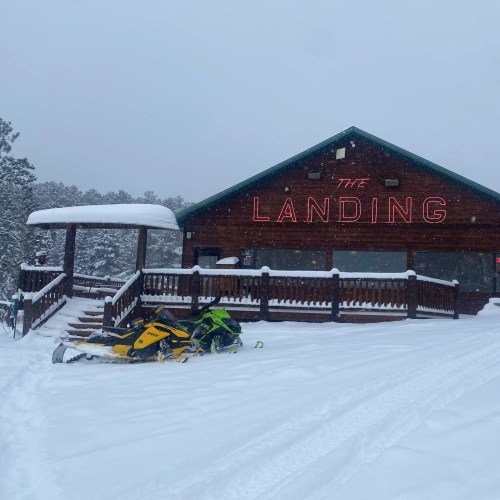 A snowy scene featuring a wooden building labeled "The Landing" and two snowmobiles parked in front.