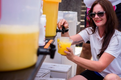 A woman in sunglasses pours a yellow drink from a dispenser into a cup at an outdoor event.