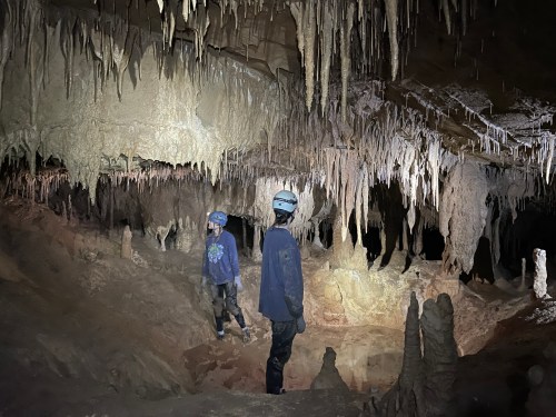 Two cavers explore a dimly lit cave, surrounded by stalactites and stalagmites, wearing helmets and headlamps.