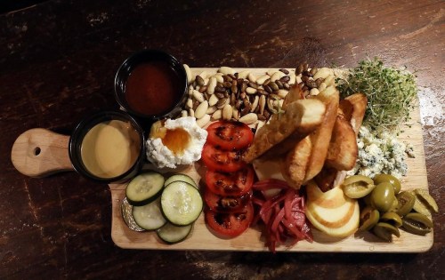 A wooden platter with assorted snacks: dips, nuts, sliced vegetables, cheese, and crispy bread.