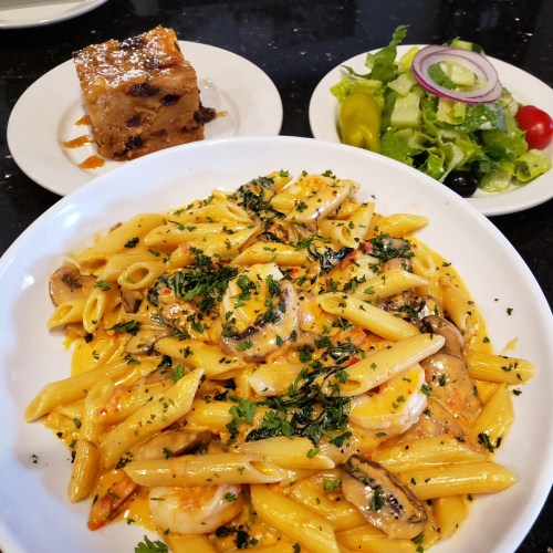 A plate of creamy shrimp penne pasta garnished with herbs, alongside a salad and a slice of dessert.