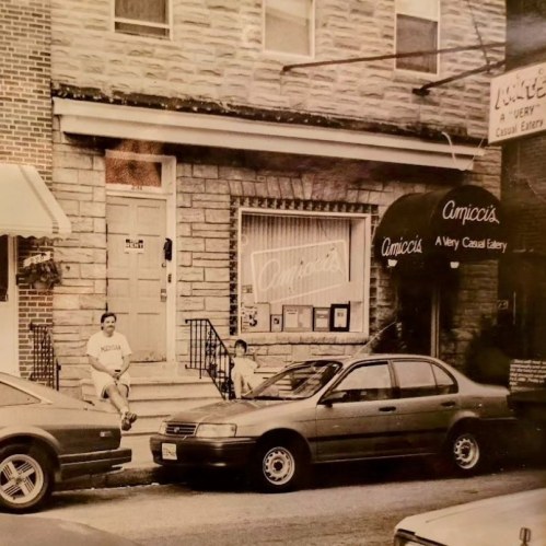 Black and white photo of a casual eatery with two people outside and parked cars on the street.