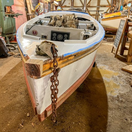A wooden boat in a workshop, featuring a white hull, blue trim, and a chain securing the front. Tools and other boats in background.