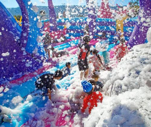 Children playing in a colorful foam-filled bounce house on a sunny day, surrounded by vibrant inflatable structures.