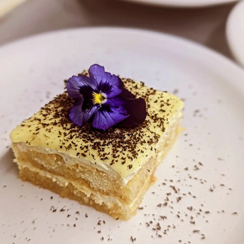 A slice of layered cake topped with yellow frosting, chocolate shavings, and a purple flower on a white plate.