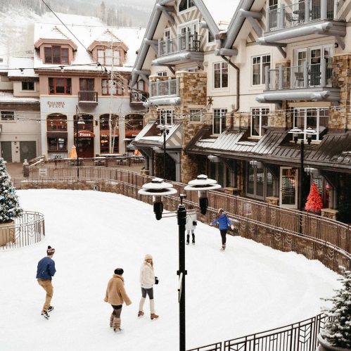 A snowy outdoor ice skating rink surrounded by charming buildings and festive decorations. People skate and enjoy the scene.