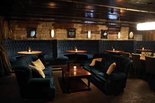 Cozy lounge with blue velvet seating, wooden tables, and soft lighting, creating an intimate atmosphere.
