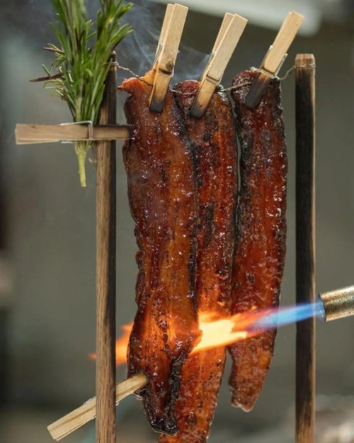 Three strips of glazed meat hanging on a wooden rack, with a flame being applied for cooking. Fresh herbs are included.