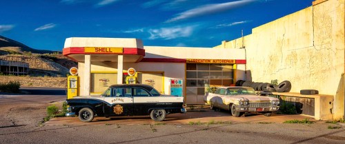 Vintage gas station with classic cars, featuring a Shell sign and an auto repair shop in a rustic setting.