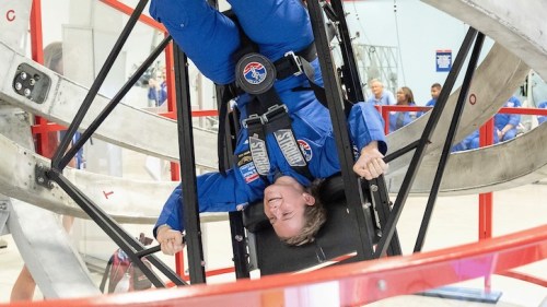 A person in a blue jumpsuit is upside down in a training simulator, smiling while strapped in.