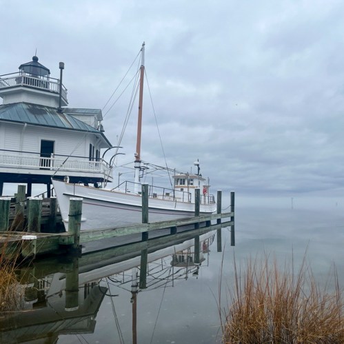 A boat docked at a pier with a white building, surrounded by calm water and a cloudy sky.