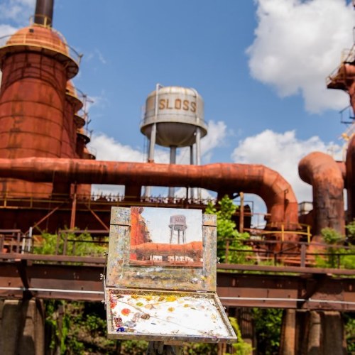 An artist's easel with a painted scene in front of an old industrial site with rusted structures and a water tower.