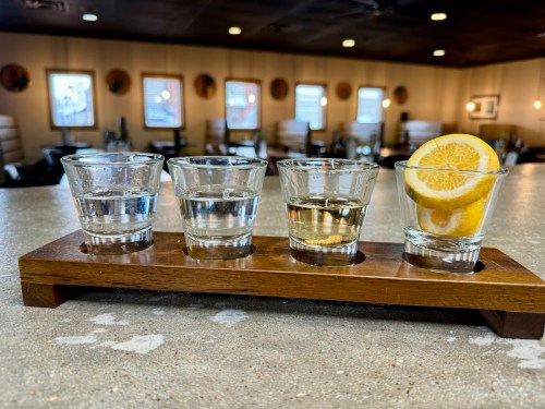 A wooden flight board holds four shot glasses with clear liquid, one featuring lemon slices, in a cozy restaurant setting.