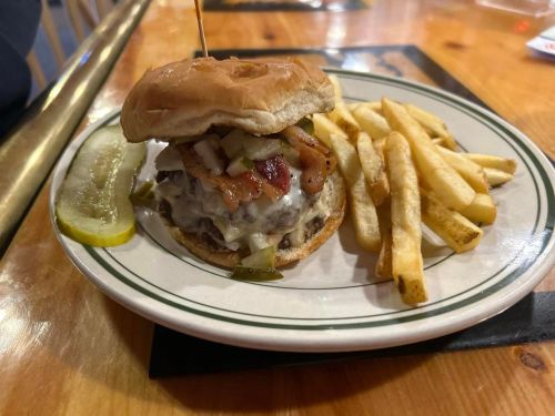 A juicy burger topped with bacon and pickles, served with a side of crispy fries and a pickle slice on a plate.