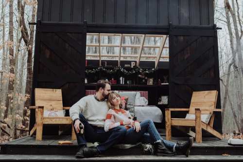 A couple relaxes together on a porch, surrounded by trees, with a cozy cabin in the background.
